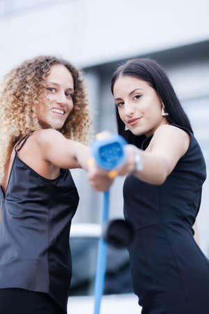 Two beautiful businesswomen in black outfut posing in front of an electric car. Girls holding blue charging cable. Shallow depth of field. Selective focus.の写真素材