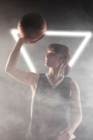 Beautiful girl with basketball. Side lit studio portrait against fog background with white neon...
