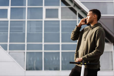 young african american man in casual outfit standing with electric scooter and talking on phone