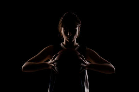Female basketball player. Beautiful girl holding ball. Side lit silhouette studio portrait against black background.の写真素材