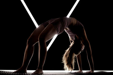Fit woman practicing yoga poses. Silhouette girl doing exercise in studio against black background with v shaped white led tube light. No stress inner balance concept.の写真素材