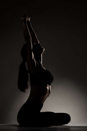 Fit girl resting and stretching after practicing yoga poses. Side lit half silhouette against dark background.の写真素材