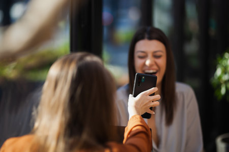 Two young business women in a cafe having one on one meeting. Friends after work talking gossiping and having coffee at a window table with reflections.の写真素材