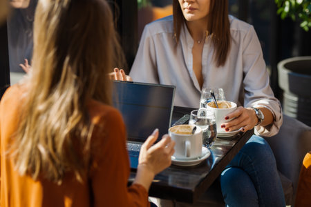 Two young business women in a cafe having one on one meeting. Friends after work talking gossiping and having coffee at a window table with reflections.の写真素材