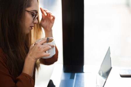 Young businesswoman in a cafe bar or restaurant. Freelancer girl working on laptop and having tea at a window table.の写真素材