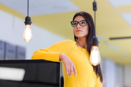 Portrait of a very beautiful brunette business woman with glasses in front of decorative light bulb in a modern office.の写真素材