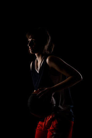 Female basketball player. Beautiful girl holding ball. Side lit half silhouette studio portrait against black background.の写真素材
