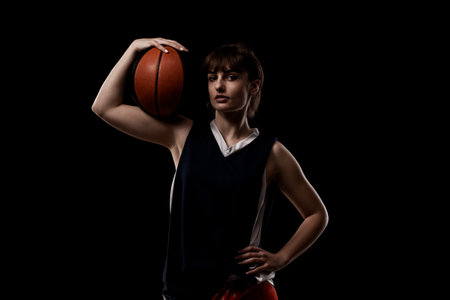 Female basketball player. Beautiful girl holding ball. Side lit half silhouette studio portrait against black background.の写真素材