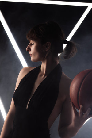 Beautiful girl in a fancy black dress holding basketball. Studio portrait against fog background with white neon lights.の写真素材