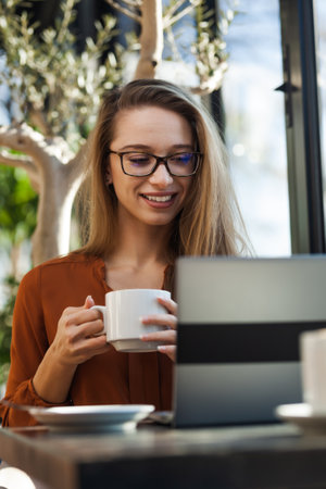 Young businesswoman in a cafe bar or restaurant. Freelancer girl working on laptop and having tea at a window table.の写真素材