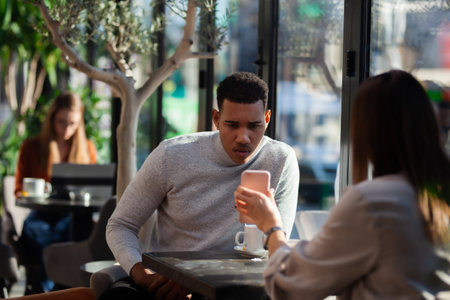 Two friends in a restaurant talking smiling and drinking tea. Multuracial business colleagues having a meeting after work or during coffee break at a cafe bar. Freelance lady working in the background.の写真素材