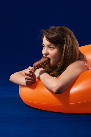 Happy girl in inflatable orange rubber ring enjoyng chocolate ice cream. Summer vacation concept. Blue surface and background.の写真素材