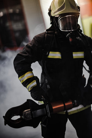 Fireman portrait wearing full equipment with oxygen mask and power hydraulic cutting tool. Smoke and fire trucks in the background.の写真素材