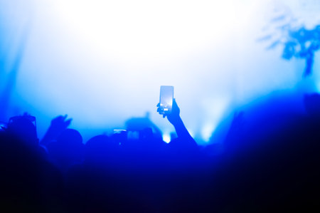Silhouette of a hand holding a smartphone capturing a moment during a night concert lit by stage lightsの写真素材