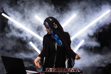 A female DJ with vibrant blue hair is actively mixing music at a turntable during a live set. Smoke and neon led tube lights in the backgroundの写真素材