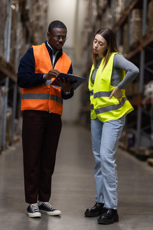 Warehouse employees collaborate using a tablet to check inventory, emphasizing teamwork and safety. They wear high visibility vests and are surrounded by shelves stacked with goods.の写真素材