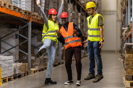 Three warehouse workers in safety gear celebrate teamwork and success in an organized storage area....