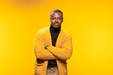 A confident man stands with folded arms, wearing a yellow cardigan against a golden background. The image exudes warmth and self-assurance in a studio environment.の写真素材