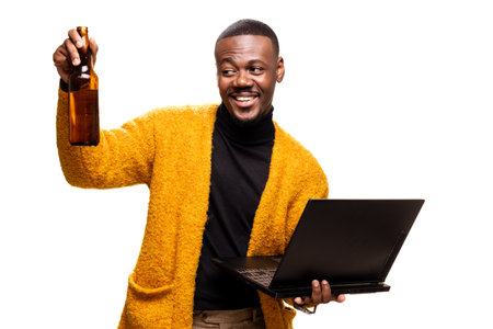 A person wearing a yellow sweater holds a beer bottle and a laptop, embodying a blend of work and relaxation.の写真素材
