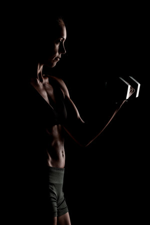 A silhouette of a muscular woman lifting a dumbbell, emphasizing strength and fitness. The artistic shadowing and dark background create a dramatic and focused image of athleticism and determination.の写真素材