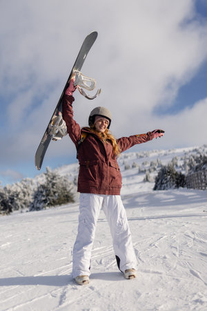 A snowboarder enthusiastically holds up their board while standing on a snowy mountainside, expressing joy for the outdoors. The background features a bright sky and frost-covered pine trees.の写真素材