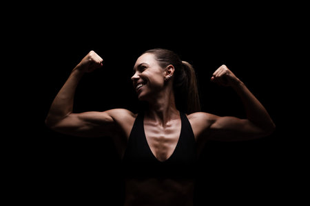 Vivid portrait of a strong and confident female athlete, showcasing her toned and muscular arms in a dramatic low-light studio environment, symbolizing empowerment, fitness, and determination.の写真素材