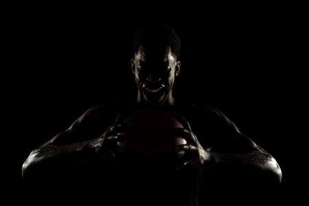 Basketball player holding a ball against black background. Distressed african american man screaming. Muscular person sidelit silhouette.の写真素材
