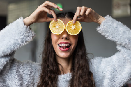 Joyful young woman with braces creatively holding lemon slices as glasses at homeの写真素材