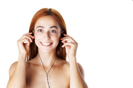 A joyful young red-haired woman wears earphones, smiling as she listens to music, isolated on a white backdrop.の写真素材