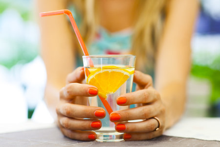 Focused close-up shot of a womans hands holding a glass with an orange slice and straw, emphasizing refreshment and a casual outdoor setting.の写真素材