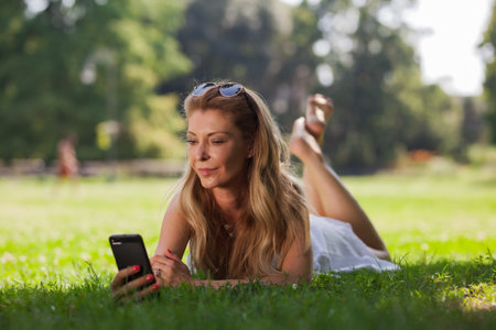 A relaxed young woman lies on the grass in a sunny park while using her smartphone, embodying leisure and connectivity.の写真素材