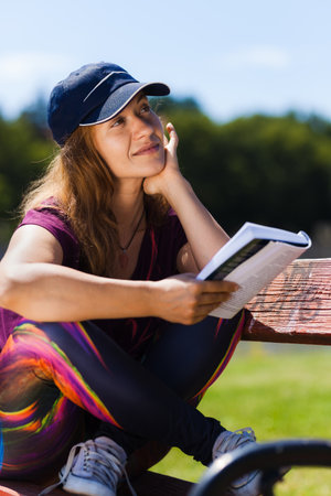 A thoughtful woman enjoys reading a book outdoors, sitting on a park bench in bright sunlight, surrounded by greenery.の写真素材