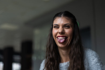 Joyful Young Woman With Braces Expressing Playfulness With a Tongue-Out Pose Indoorsの写真素材
