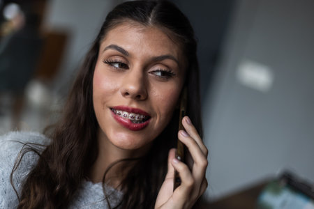 Smiling Young Woman with Braces Talking on a Phone Indoorsの写真素材