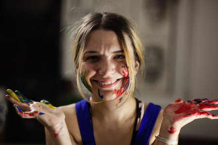 A cheerful female painter shows her hands covered in vibrant paint, her face smeared with colors,...