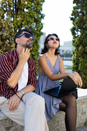 Casual outdoor meeting with a man and woman in sunglasses looking thoughtful with a laptop on a sunny dayの写真素材