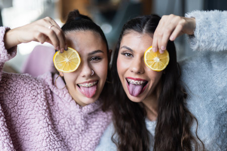 Two joyful young women posing playfully with lemon slices, enjoying a lighthearted moment together indoorsの写真素材