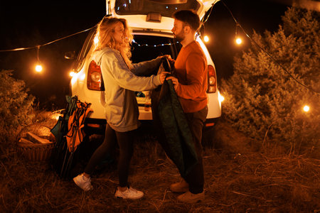 Couple enjoys cozy evening while setting up a tent under string lights near their car in nature...
