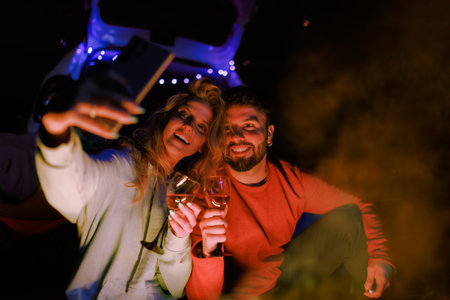 Couple enjoys a cozy evening by the campfire while taking selfies, surrounded by nature during a...
