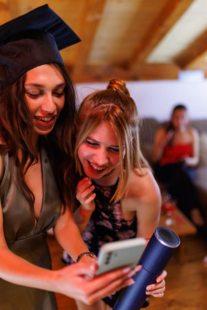 Two young women celebrating graduation together, sharing a joyful moment viewing a smartphone inside...