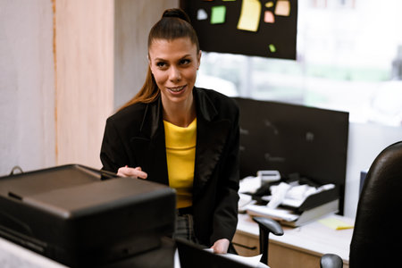 Office setting with a woman in a black blazer smiling while holding documents near a printer during midday work hoursの写真素材