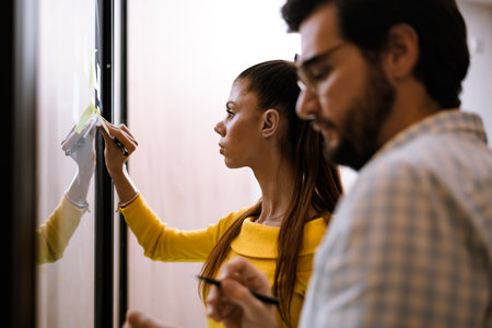 Two coworkers collaborating at a modern office space, brainstorming ideas while writing on a glass board during a sunny afternoonの写真素材