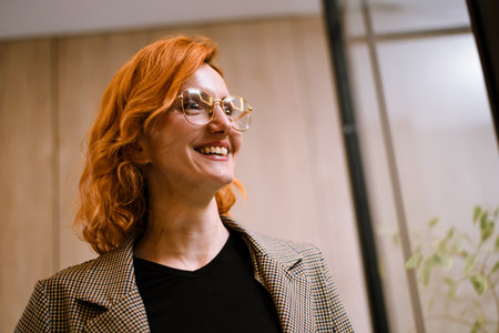 Smiling woman with orange hair and glasses in a modern indoor setting, enjoying her surroundings at a casual event in the eveningの写真素材