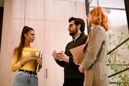 Group discussion in modern office space with two women and a man engaged in conversation while holding notebooksの写真素材