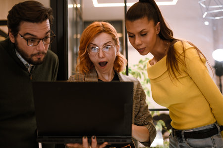 Friends gather around a laptop in an indoor setting, sharing excitement and surprise as they review something on the screen together during a casual eveningの写真素材