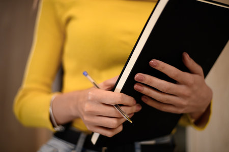 Woman in a yellow sweater writing notes with a pen in a notebook at home during the day while preparing for an important projectの写真素材