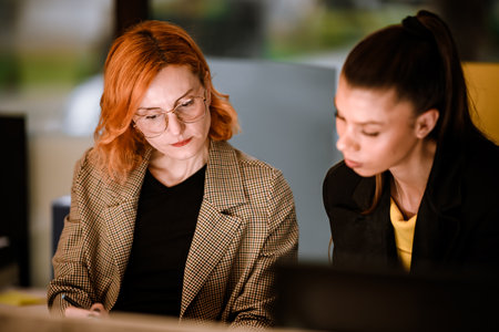 Two women collaborating on a project in a modern office during evening hours, showcasing teamwork and focused discussion on work tasksの写真素材