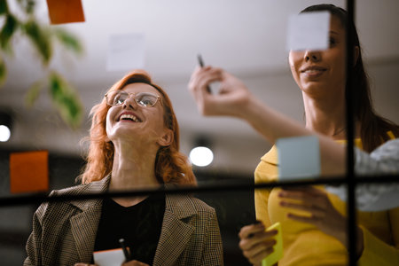 Creative brainstorming session with two women collaborating in a modern office space filled with colorful sticky notes on a glass wall during a productive afternoonの写真素材