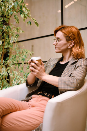 Woman with orange hair enjoying coffee in a modern cafe setting surrounded by plants in the afternoon lightの写真素材