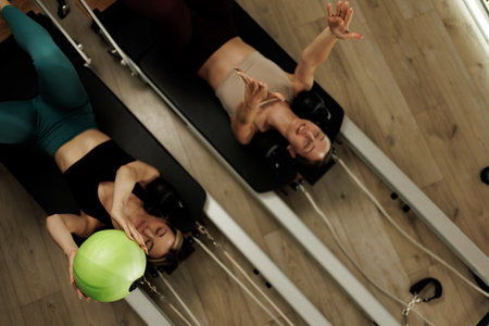 Two women engaged in a reformer pilates workout in a sunlit studio focusing on strength training and flexibility in the early morningの写真素材